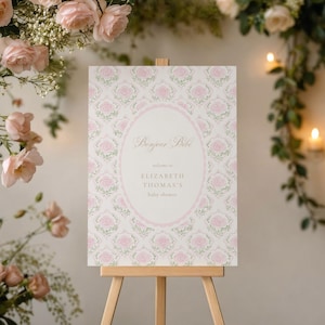 May include: A baby shower sign on a wooden easel. The sign features a floral pattern with pink roses and the text "Bonjour Bébé." The sign also includes the text "Welcome to Elizabeth Thomas's baby shower."