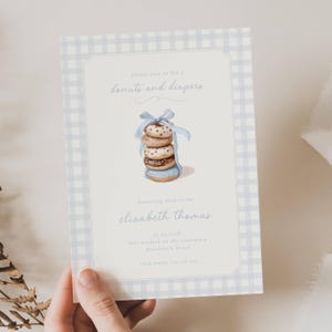 Może przedstawiać: Zaproszenie na baby shower z jasnoniebieską obwódką w kratkę. Zaproszenie zawiera stos akwarelowych pączków związanych niebieską wstążką. Tekst zawiera "donuts and diapers".