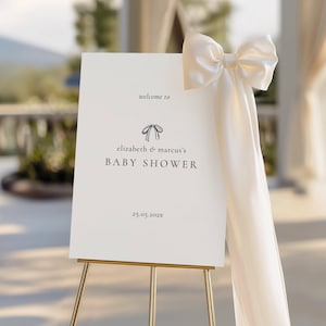 May include: A white baby shower welcome sign on a gold easel. The sign reads "welcome to" above "elizabeth & marcus's BABY SHOWER" and the date "25.05.202x". A cream-colored bow and fabric are draped on the sign.