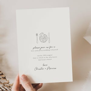 以下が含まれることがあります： クリーム色の結婚式の招待状。ピザのスライスのグラフィック、フォーク、ナイフが描かれています。「please join us for a love and pizza wedding rehearsal」というテキストと日付、時間、場所が記載されています。「Christie + Marcus」の名前が下にあります。