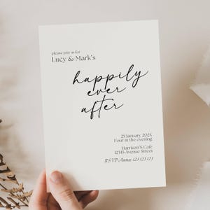May include: A cream-colored invitation with black script that reads "please join us for Lucy & Mark's happily ever after." Additional text includes the date, time, location, and RSVP information. The invitation is held by a hand.