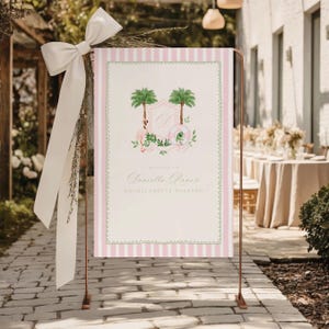 Op de afbeelding: Een welkomstbord voor een vrijgezellenfeest, met een roze en witte gestreepte achtergrond. Het bord toont een bloemen- en palmbomenillustratie met de tekst "Welcome to Daniella's Bachelorette Weekend". Het wordt ondersteund door een koperkleurige metalen standaard en een witte strik.
