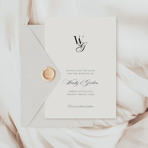 Könnte beinhalten: Eine cremefarbene Save-the-Date-Hochzeitskarte mit passendem Umschlag, verschlossen mit einem goldenen Wachssiegel. Die Karte zeigt das Monogramm WG, die Namen Wendy und Gordon, das Datum 25. Juli 2029 und den Text Formal invitation to follow.