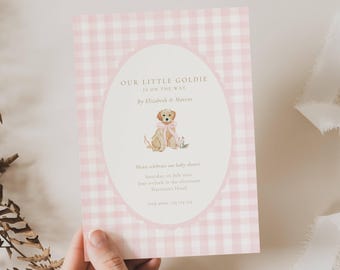 May include: A baby shower invitation with a pink gingham background. The invitation features the text "OUR LITTLE GOLDIE IS ON THE WAY" and a watercolor illustration of a golden retriever puppy wearing a pink bow. The invitation is held by a hand.