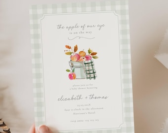 May include: A baby shower invitation with a watercolor illustration of apples in a metal bucket, a green and white checkered towel, and text. The invitation has a green and white gingham border and reads "the apple of our eye is on the way."