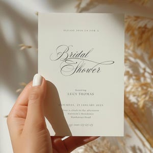 May include: A white card with black text announcing a bridal shower for Lucy Thomas. The shower will be held on Saturday, January 25th at four o'clock in the afternoon at Harrison's Residence on Manhattan Road.