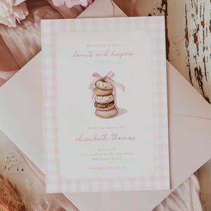Może przedstawiać: Różowo-białe zaproszenie na baby shower w kratkę z napisem "donuts and diapers". Zaproszenie przedstawia stos pączków związanych różową wstążką. Zaproszenie jest w różowej kopercie.