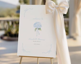 May include: A welcome sign for a bridal shower, displayed on a gold easel. The sign is white with light blue floral accents and text that reads "welcome to Elizabeth Thomas Bridal Shower 23.03.202X." A large cream-colored bow adorns the side.