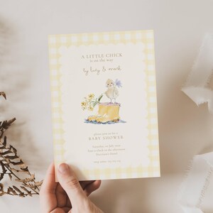May include: A baby shower invitation with a yellow gingham border. The invitation features a watercolour illustration of a chick standing in a yellow boot, holding flowers. The text reads "A LITTLE CHICK is on the way" and invites guests to a baby shower.