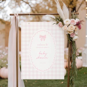 May include: A baby shower sign with a pink gingham background and a scalloped oval frame. The frame contains the text "welcome to elizabeth dane's baby shower" and a bow illustration. The sign is decorated with flowers and ribbons.