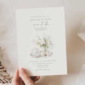 May include: A white invitation card with a watercolor illustration of a teacup, teapot, and macarons on a silver tray. The text on the card reads "Please join us for Time for Tea with mom-to-be Elizabeth Thomson Saturday, 25 January 2025 four o'clock in the afternoon St. James Chapel Manhattan Road 123 123 123".