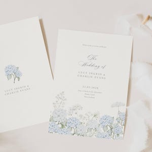 May include: Two ivory wedding invitations with blue hydrangea watercolor floral accents. One invitation features a single floral cluster, while the other displays the text "The Wedding of LUCY INGRID & CHARLIE EVANS" and event details.