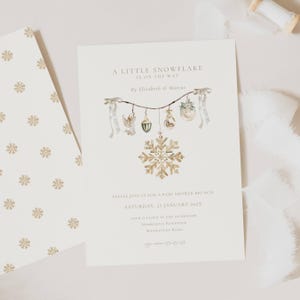 May include: A baby shower invitation with the text "A Little Snowflake Is On The Way." The invitation features a gold snowflake design and watercolor illustrations of ornaments. The invitation is on a cream background with a matching envelope.