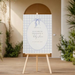 May include: A baby shower welcome sign on a wooden easel. The sign features a light blue gingham pattern background with an oval in the center. The oval contains the text "Welcome to Elizabeth Thomson's baby shower" and the date "25 January 2023".
