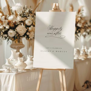 May include: A cream-colored wedding sign on a wooden easel. The sign reads "Happily ever after" in elegant script, with "Let's celebrate Claire & Marcus" below. The background features floral arrangements and decorative vases.