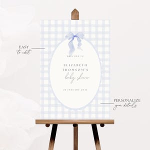 May include: A baby shower sign on a wooden easel. The sign has a light blue gingham pattern background with an oval in the center. The oval contains the text "WELCOME TO ELIZABETH THOMSON'S baby shower" and the date "25 JANUARY 2025".