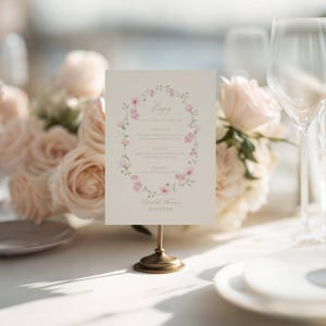 May include: A cream-colored menu card with a floral wreath design, held by a gold-toned stand. The menu includes the word "Enjoy" at the top, with sections for appetizers, entree, and dessert. The bottom of the card reads "Elizabeth & Samuel's Baptism".