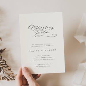 May include: A cream-colored wedding invitation with elegant script that reads "Nothing fancy, Just love." The invitation includes details for the wedding of Claire & Marcus, including the date, time, and location.