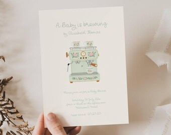 May include: Baby shower invitation featuring a mint green espresso machine illustration with floral accents. The text "A Baby is brewing" and event details are displayed on the card. The invitation is held by a hand.