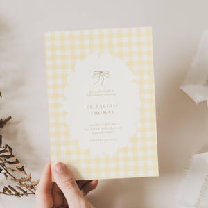 Può includere: Un invito per un baby shower con sfondo a quadretti gialli. L'invito presenta una forma ovale smerlata con un fiocco e il testo "ELIZABETH THOMAS". Sono inclusi anche la data, l'ora e il luogo.