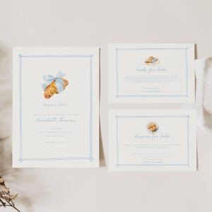 May include: Set of three baby shower invitations. The invitations are light cream with a blue border and watercolor illustrations of croissants. The main invitation says "bonjour bebe!" and includes event details.