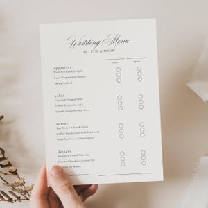 May include: A cream-coloured wedding menu card featuring the text "Wedding Menu by LUCY & MARK" in a decorative font. The menu lists appetizers, salads, entrees, and desserts, with spaces for names and check boxes. The card is held by a hand.