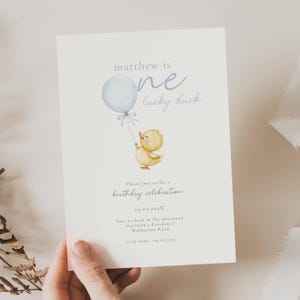 Puede incluir: Una invitación de cumpleaños crema con una ilustración de un patito amarillo sosteniendo un globo azul. El texto dice "matthew is one lucky duck" y "birthday celebration". La invitación incluye la fecha, hora y lugar.