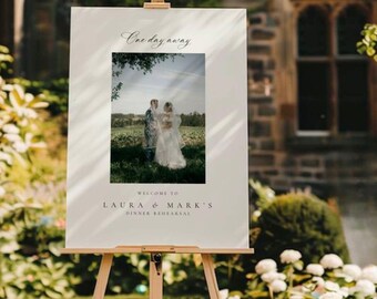 Könnte beinhalten: Ein weißes Hochzeits-Willkommensschild auf einer Holzstaffelei. Das Schild zeigt ein Foto eines Paares und den Text "One day away". Unter dem Foto steht "Welcome to Laura & Mark's Dinner Rehearsal."