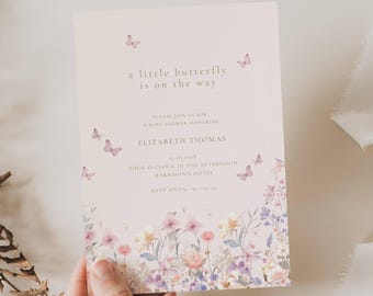 May include: A baby shower invitation with a floral design and the text "a little butterfly is on the way." The invitation features a soft pink background with watercolor flowers and butterflies. The invitation is held by a hand.