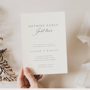 May include: A cream wedding invitation with the text "NOTHING FANCY Just love." The invitation includes the wedding details: date, time, and location. The names "CLAIRE & MARCUS" are also featured. The invitation is held by a hand.