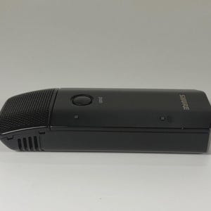 May include: A black Shure wireless microphone transmitter with a rectangular body, a textured grille on the front end, a circular power button labeled push, and small indicator lights along the side of the smooth, matte plastic casing.