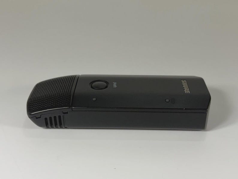 May include: A black Shure wireless microphone transmitter with a rectangular body, a textured grille on the front end, a circular power button labeled push, and small indicator lights along the side of the smooth, matte plastic casing.