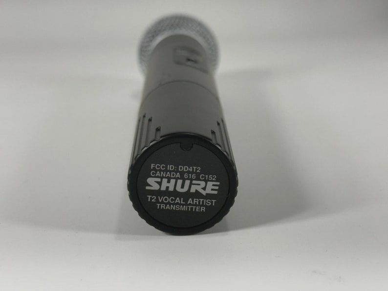 Puede incluir: Un micr&oacute;fono Shure T2 Vocal Artist Transmitter gris. El micr&oacute;fono tiene una cabeza de malla plateada y un cuerpo gris oscuro. La parte inferior del micr&oacute;fono tiene el logotipo de Shure y el texto "T2 Vocal Artist Transmitter".
