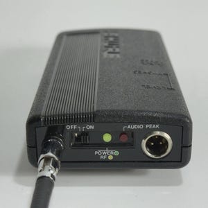 Puede incluir: Un dispositivo de audio negro con una superficie texturizada y un cable conectado. El dispositivo tiene un interruptor de encendido/apagado, indicadores de alimentaci&oacute;n y RF, y un puerto de pico de audio. La palabra "Clarion" es visible.