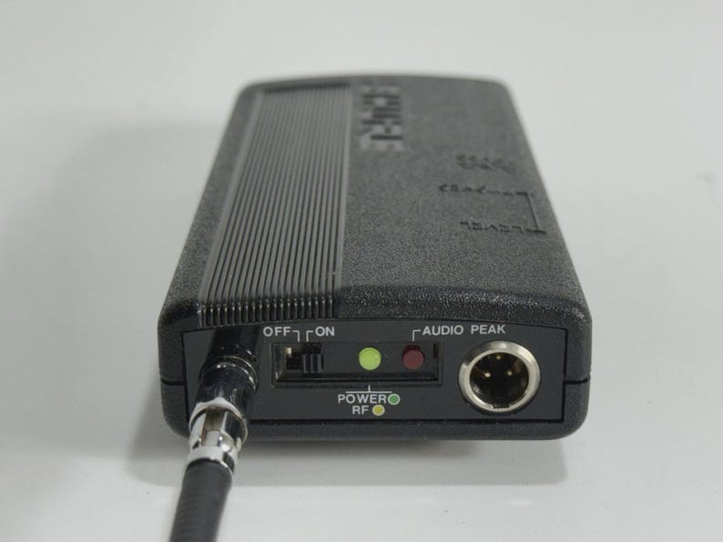 Puede incluir: Un dispositivo de audio negro con una superficie texturizada y un cable conectado. El dispositivo tiene un interruptor de encendido/apagado, indicadores de alimentaci&oacute;n y RF, y un puerto de pico de audio. La palabra "Clarion" es visible.