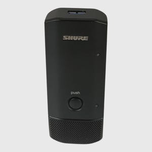 May include: A black Shure wireless microphone bodypack transmitter featuring a top-mounted USB port, a central circular push button labeled with the word push, and a perforated speaker-style grille section at the bottom of the rectangular unit.