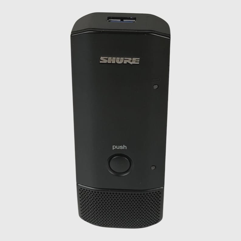 May include: A black Shure wireless microphone bodypack transmitter featuring a top-mounted USB port, a central circular push button labeled with the word push, and a perforated speaker-style grille section at the bottom of the rectangular unit.