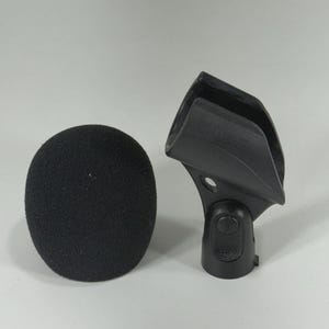 Puede incluir: Un filtro antipop negro y un soporte negro para micr&oacute;fono. El filtro antipop tiene forma ovalada y redondeada, hecho de un material poroso. El soporte es de pl&aacute;stico duro y tiene la marca "SHURE".