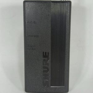 Puede incluir: Un dispositivo de audio rectangular negro con la marca "SHURE" impresa verticalmente. El dispositivo tiene una antena plateada y texto que indica "LEVEL" y "PHONES". Probablemente se utiliza para la transmisi&oacute;n o recepci&oacute;n de audio.