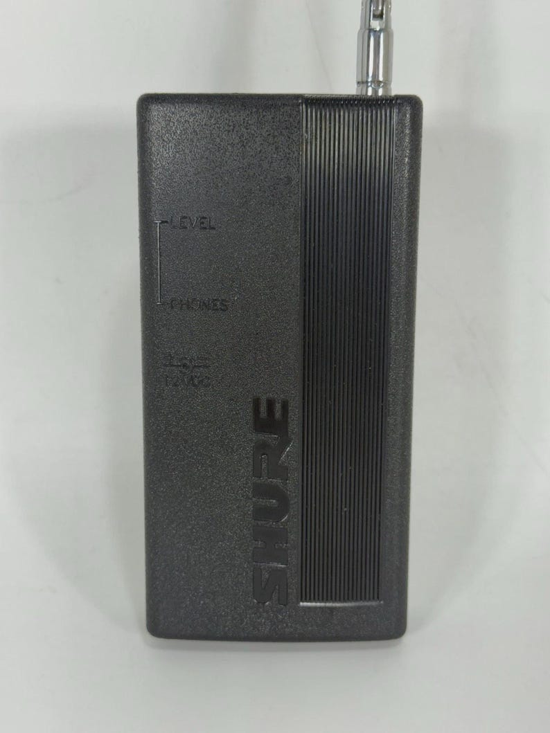Puede incluir: Un dispositivo de audio rectangular negro con la marca "SHURE" impresa verticalmente. El dispositivo tiene una antena plateada y texto que indica "LEVEL" y "PHONES". Probablemente se utiliza para la transmisi&oacute;n o recepci&oacute;n de audio.