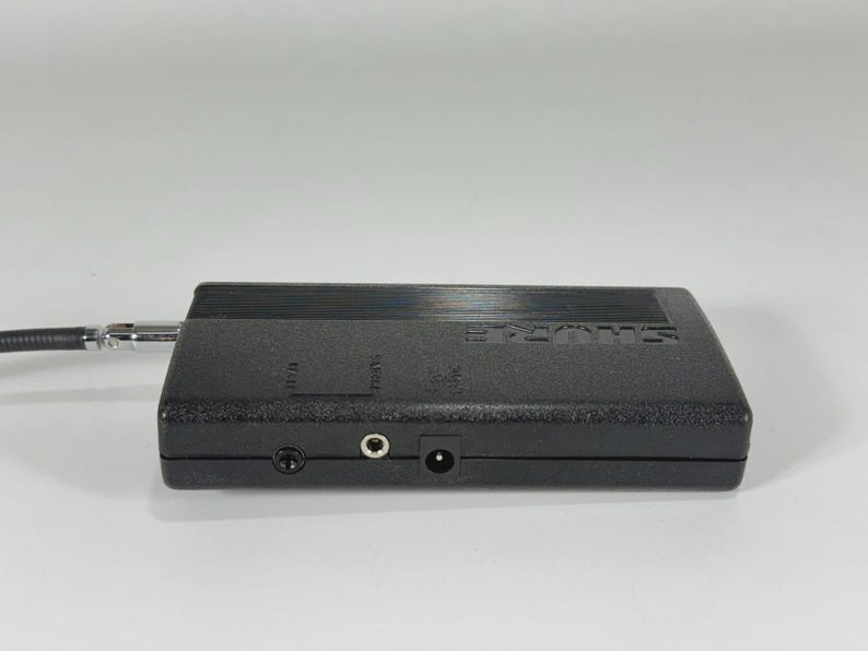 Puede incluir: Un dispositivo de audio negro Shure con un cable adjunto. El dispositivo tiene varios conectores de entrada y el logotipo de Shure. El dispositivo es rectangular y tiene una superficie texturizada.