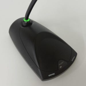 May include: A black Shure Microflex boundary microphone with a triangular base, a push button, and a green LED indicator ring at the base of the cable. The device features the brand name Shure and the word Microflex printed on the top surface.