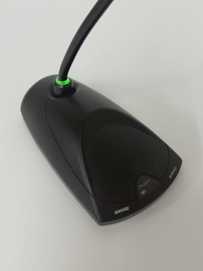 May include: A black Shure Microflex boundary microphone with a triangular base, a push button, and a green LED indicator ring at the base of the cable. The device features the brand name Shure and the word Microflex printed on the top surface.