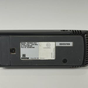 May include: A black Shure MXW6/C Z10 wireless microphone transmitter with a rectangular base. The label displays technical specifications including frequency 1920-1930 MHz, IC 616A-MXW6, FCC ID DD4MXW6, input 5V 500mA, and serial number 3QH2537659.