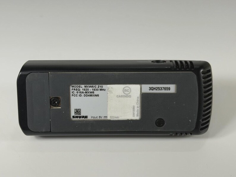 May include: A black Shure MXW6/C Z10 wireless microphone transmitter with a rectangular base. The label displays technical specifications including frequency 1920-1930 MHz, IC 616A-MXW6, FCC ID DD4MXW6, input 5V 500mA, and serial number 3QH2537659.