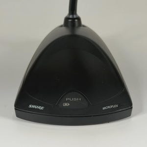 May include: A black Shure Microflex boundary microphone base with a triangular shape. The front panel features a central button labelled PUSH with a battery icon, the brand name SHURE on the left, and the word MICROFLEX on the right side.