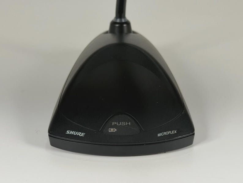 May include: A black Shure Microflex boundary microphone base with a triangular shape. The front panel features a central button labelled PUSH with a battery icon, the brand name SHURE on the left, and the word MICROFLEX on the right side.