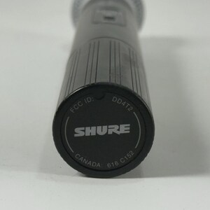 May include: Close-up of a black Shure microphone. The base of the microphone is visible, displaying the brand name "SHURE" in white, along with "FCC ID", "DD4T2", "CANADA", and other markings. The microphone has a textured, cylindrical body.
