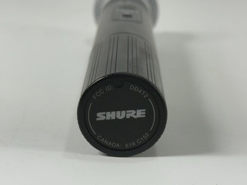 May include: Close-up of a black Shure microphone. The base of the microphone is visible, displaying the brand name "SHURE" in white, along with "FCC ID", "DD4T2", "CANADA", and other markings. The microphone has a textured, cylindrical body.