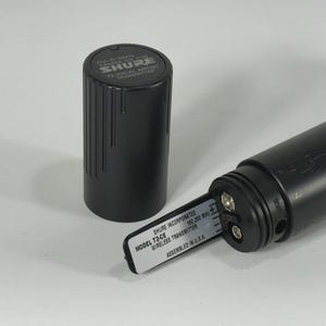 Puede incluir: Transmisor de micr&oacute;fono inal&aacute;mbrico Shure T2-CE negro con un compartimento de bater&iacute;a cil&iacute;ndrico negro y una tapa negra separada. El transmisor tiene una etiqueta que dice "MODEL T2-CE WIRELESS TRANSMITTER ASSEMBLED IN U.S.A."